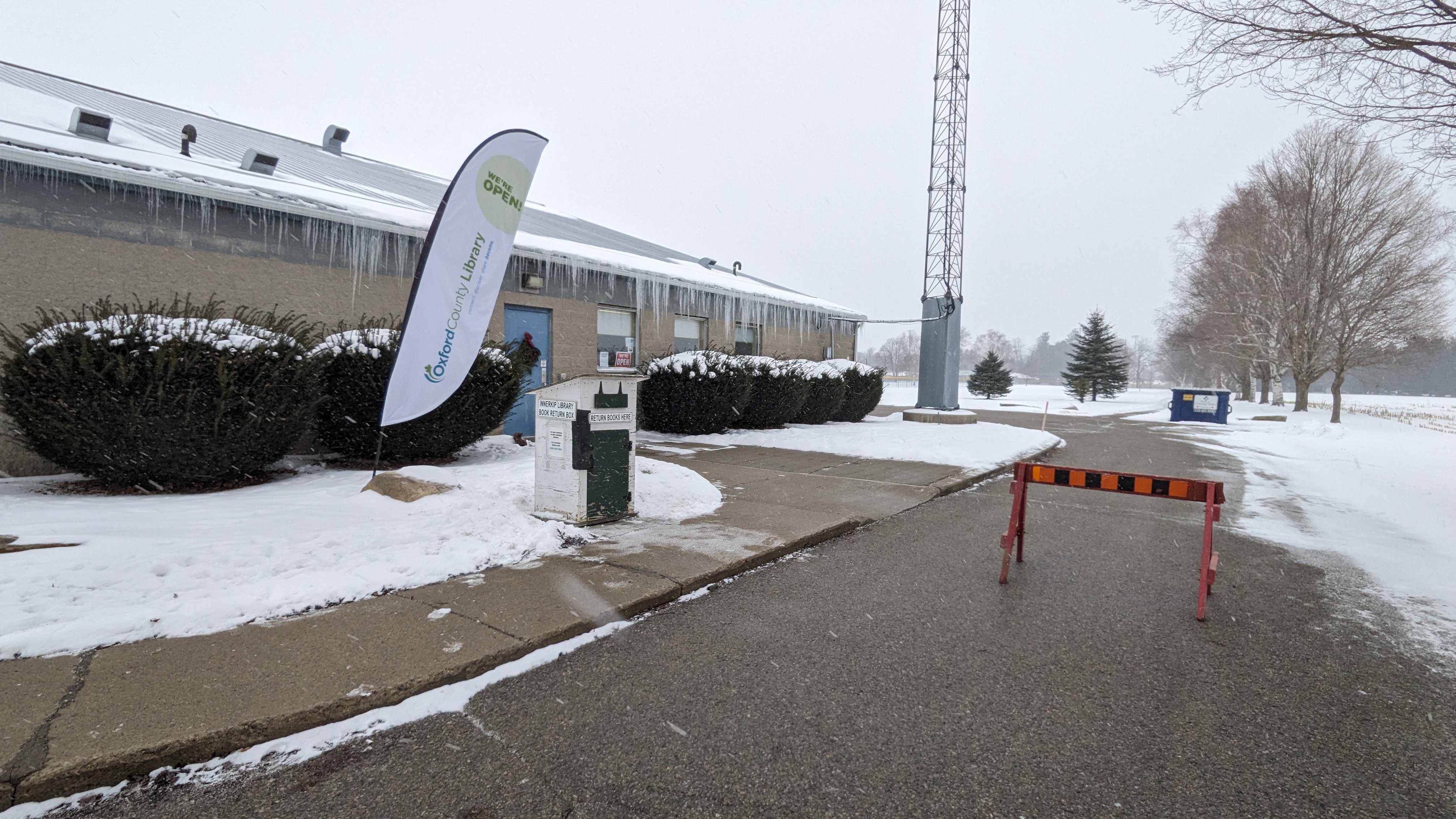 Snowy library building with icicles, a "We're Open" flag, a book return box, and a barricaded driveway.