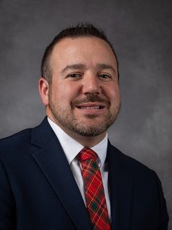 Councilor Brian Petrie wearing a dark blue suit with a red, blue, green and yellow plaid tye, he has short dark brown hair. 