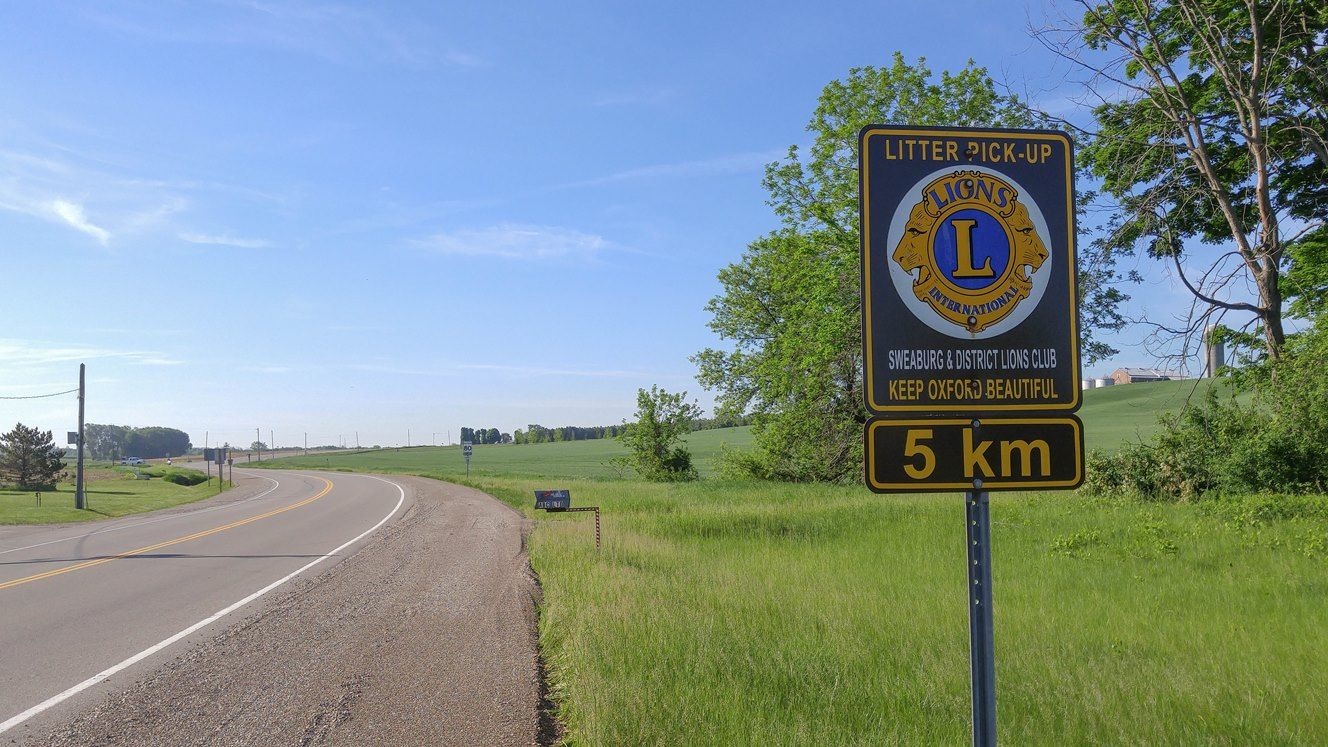 road bending to the left, with grassy area on the side and a few trees with a sign that states the litter is picked up by the Lions group for the next 5km.